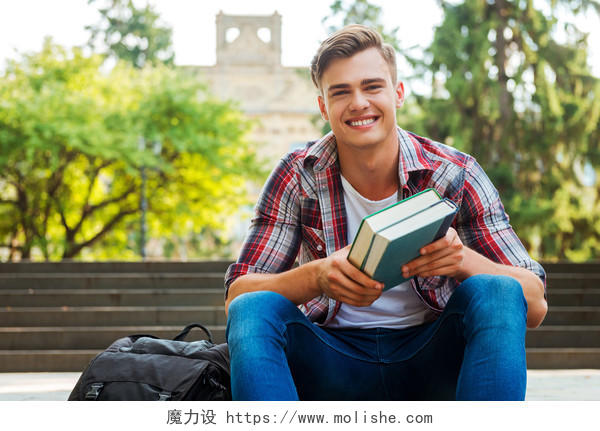 快乐的书呆子英俊的男学生拿着课本和微笑着坐在户外楼梯与高校建筑的背景